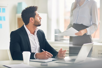 Boss in formalwear sitting by desk and talking to his secretary with papers