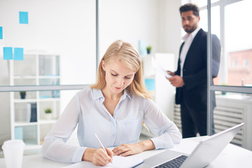 Busy young woman making notes in notebook on background of male employee