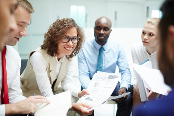 Obraz premium Happy economist with financial papers looking at camera while making report among her colleagues at meeting or seminar