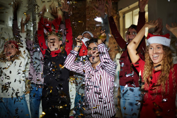 Excited young friendly people with raised hands dancing at xmas party in confetti fall