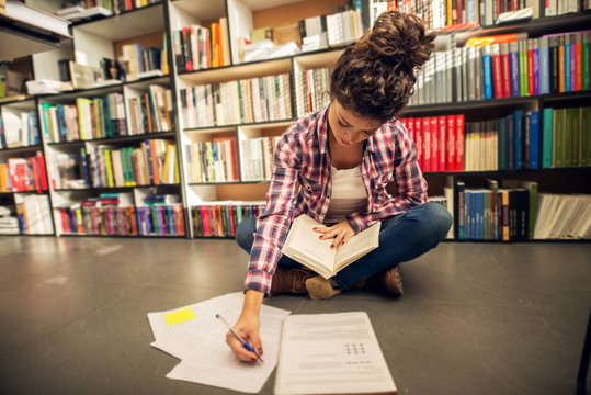 Concept of education, library, students. Close up of focused adorable curly high school student girl sitting on the floor with a book in the library in front the bookshelf and taking learning notes.