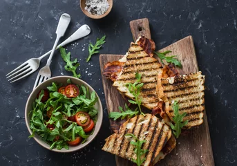 Zelfklevend Fotobehang Grilled bacon, mozzarella sandwiches on wooden cutting boards and arugula, cherry tomato salad on dark background, top view.Delicious breakfast or snack, flat lay © okkijan2010