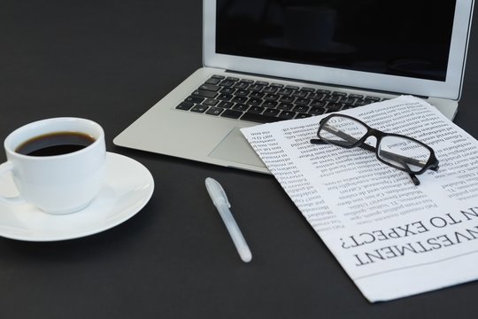 Cup of coffee, laptop, spectacles, newspaper and pen on black