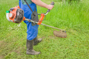 Man wearing overalls and rubber boots mowing green grass with gasoline lawn trimmer. The...