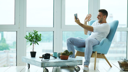 Young happy man have online video chat using digital tablet computer sitting on balcony in modern...