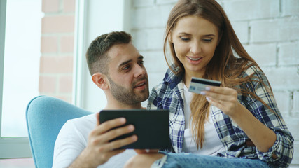 Young cheerful couple using digital tablet computer for online shopping sit on balcony in modern loft apartment