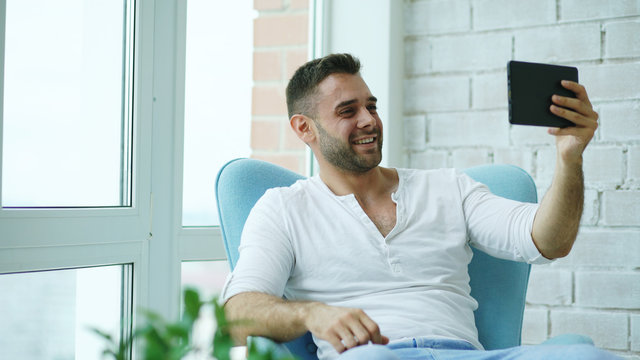 Young happy man have online video chat using digital tablet computer sitting on balcony in modern loft apartment