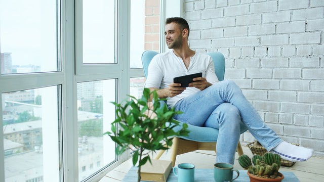 Attractive Man Using Digital Tablet Sitting In Chair At Balcony In Loft Modern Apartment