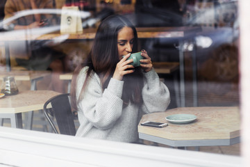portrait of young beautiful cute brunette girl in grey sweater drinking coffee at a table in  cafe throuth glass window