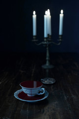 White tea cup in dark blood on dark wooden table with burning candles in candleholder.