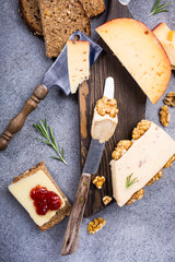 Various types of cheese with wholegrain bread, strawberry jam and walnuts on wooden board on gray stone background. Top view, flat lay.