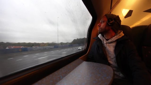 Man with headphones is riding a train and looking out the window at cars on road