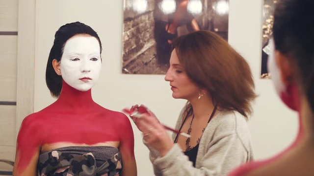 Face art. Makeup artist painting on the model's neck and shoulders with red color foudation. Reflection in the mirror