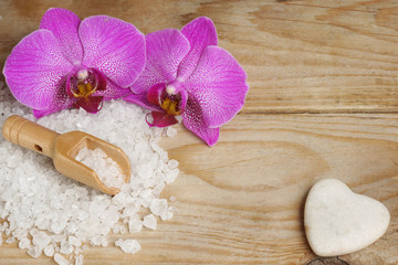 Bright orchid flowers next to white bath salt and a heart shaped stone on a wooden table