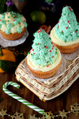 Cupcakes decorated with cream forming Christmas fir trees on a wicker basket surrounded by festive attributes on a wooden table