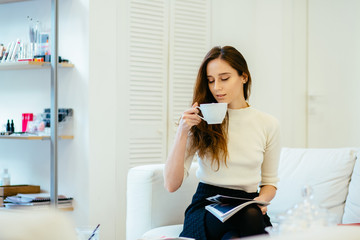 Charming stylish female client with magazine in hands drinking cup of cappuccino and waiting for make up artist at modern interior beauty parlor.