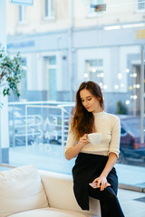 Vertical portrait of charming stylish female client with smartphone hands drinking cup of cappuccino and waiting for make up artist at modern interior beauty parlor or cafe.