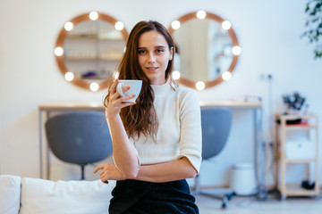 Stylish female make up artist drinking cup of cappuccino and waiting for visitors at modern interior beauty parlor.