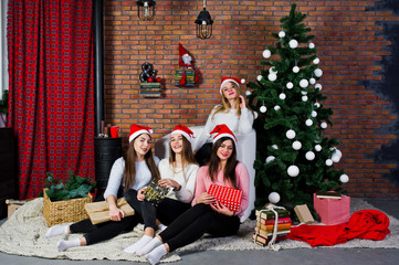 Four cute friends girls wear on warm sweaters, black pants and santa hats against new year tree with christmas decoration at studio.