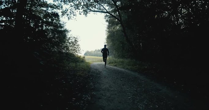 Sportsman running on dark autumn forest road. Young athlete on quiet autumn deserted pass. Cinematic drone back view.