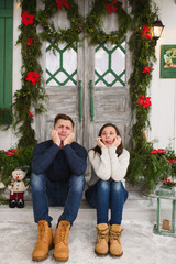 Obraz na płótnie Canvas Happy funny young couple in love dressed in sweater sitting on porch steps at light house with decorated in red green New Year door at home. Christmas good mood. Family and holiday 2018 concept