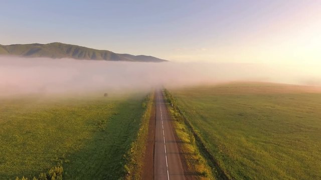 AERIAL:  An empty road in a dense fog against a backdrop of a mountain landscape. Beautiful nature during sunrise. Highway, transportation, outdoor, road trip.