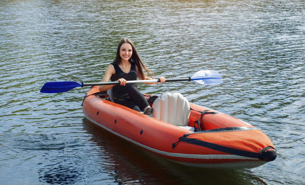 The smiling girl -the sportswoman with longdark hair in blacksportswear rows with an oar on the lake in a red inflatable canoe in a warm summer sunny day. Occupation by sports rowing on a kayaking.