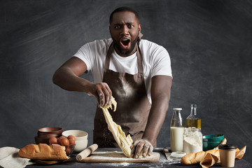 Shocked dark skinned professional male baker prepares dough at kitchen, kneads dough, can`t understand why it is sticky, needs more flour to make thick, isolated over black chalk background. - obrazy, fototapety, plakaty