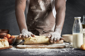 Image of hard working male cook or baker with dark skin wears apron, sheets well made dough, going...