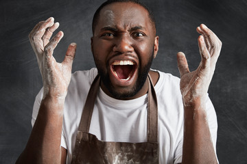 Headshot of furious dark skinned male chef yells with anger at cooks who spoiled dish for client or permanent visitor of cafe, gestures with hands, expresses aggression. Negative emotions concept