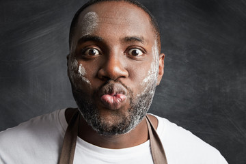 Close up portrait of funny male with dark skin and dirty face, rounds lips and makes grimace, foolishes as prepares and cooked everything, isolated over chalk black background. Facial expressions