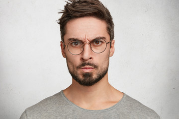 Headshot of strict annoyed aggressive man with beard, mustache, looks seriously through round spectacles, frowns face in dissatisfaction, expresses negative emotions, isolated over white concrete wall