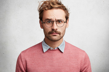 Fototapeta premium Portrait of attractive bearded young male model looks in stupor and dissatisfaction directly into camera, shocked to read news or have conversation with interlocutor, poses against white background