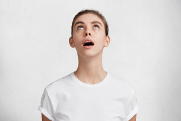 Curious beautiful woman looks with intriguing expression upwards, notices something interesting, keeps mouth opened, dressed in white casual t shirt, isolated over concrete wall. Curiosity concept