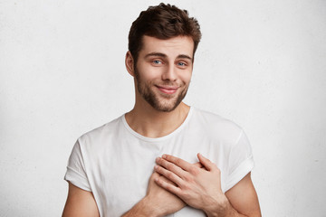 Kind pleasant looking good natured male with appealing appearance looks with pleased expression into camera, keeps hands on heart, expresses his positive feelings towards girlfriend, isolated on white