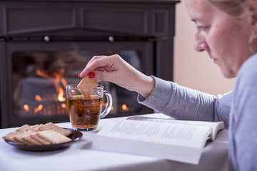 A woman drinks tea and reads a book in front of the fireplace