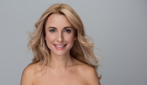Beauty concept. Portrait of happy attractive middle-aged woman is standing with naked shoulders and looking at camera with wide smile. Isolated background with copy space in the right side