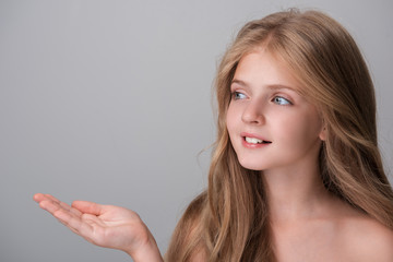 Look here. Joyful pretty girl with long hair is looking aside with smile while holding out her hand palm up. Isolated background with copy space in the left side