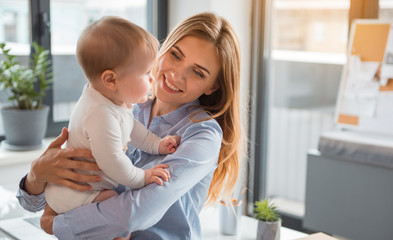 Portrait of cheerful mom keeping serene child on hand in modern room. Infant at job