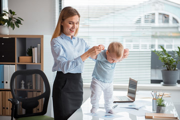 Fototapeta premium Portrait of laughing woman keeping full length interested baby by arms. He standing on table in office. Job, fun and child concept