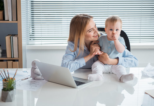 Portrait of smiling mother telling with calm kid. He keeping finger in mouth while locating on desk. Occupation and family concept