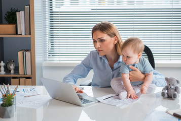 Portrait of serene businesswoman typing in laptop. interested baby looking at it while locating on desk in apartment. Labor and family concept