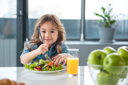 Portrait of pretty little female child having healthy breakfast. She is holding fork under the chopped vegetables and smiling