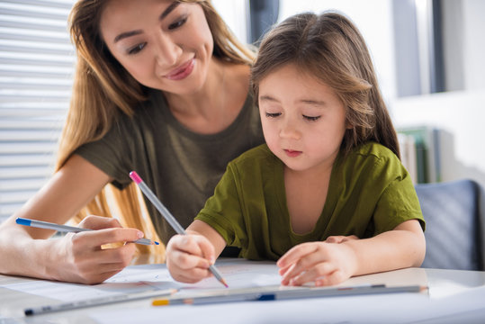 Portrait of joyful girl drawing picture with her mother with interest. She is holding pink pencil while sitting at table at home