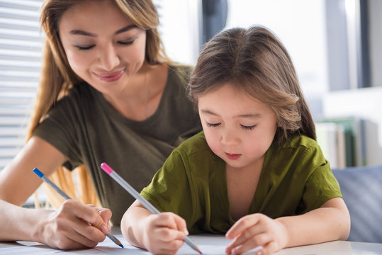 Portrait of interested asian girl painting image with her mother. They are holding colorful pencils with joy