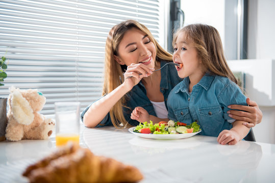 Portrait of happy asian woman feeding her little daughter by fresh vegetables. Girl is tasting tomato with joy. Family is sitting at table in kitchen