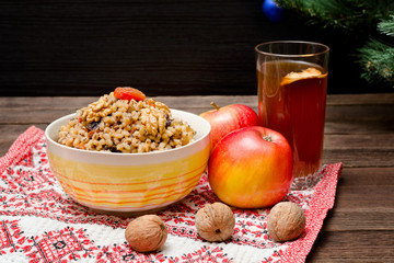Dish of traditional Slavic treat on Christmas Eve. Christmas tree, apples, walnuts, glass of compote on a patterned tablecloth. Brown and blak wooden background.