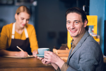 Portrait of happy young man tasting cup of delicious coffee in cafe. Smiling worker writing information on background. Rest concept