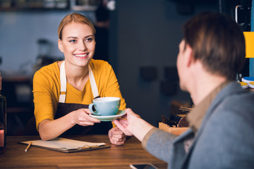 Portrait of beaming female barista providing mug of appetizing beverage to client in confectionary shop. Occupation concept