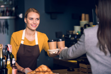Portrait of cheerful female barista giving two cups of delicious coffee to visitor. She standing at counter in cozy confectionary shop. Work concept